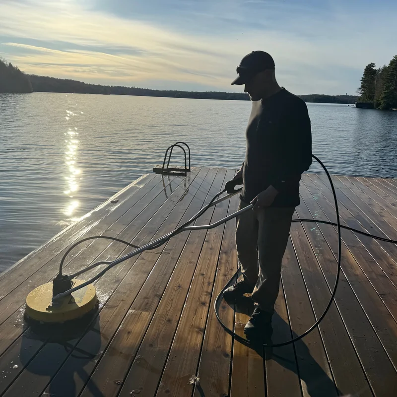 Professional deck cleaning technician using surface cleaner on Muskoka waterfront dock at sunset